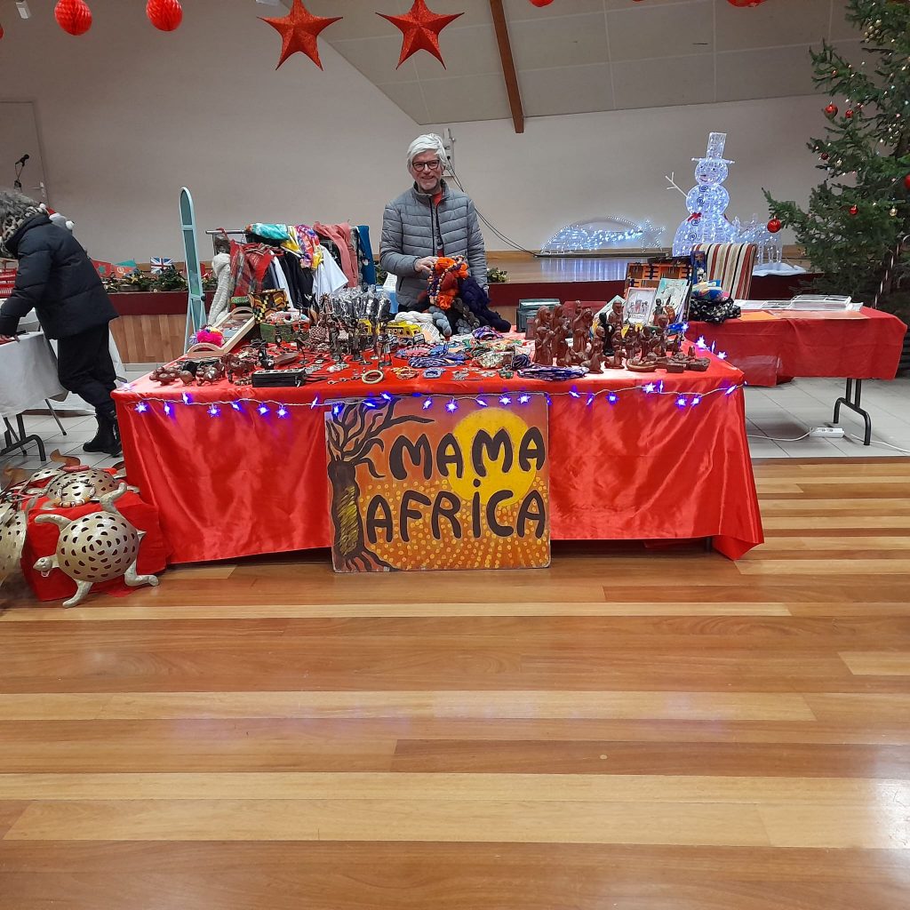 Le stand du marché de Noël de l'association humanitaire, un volontaire souriant se tenant derrière. Couverte de satin route, la table est recouverte d'une multitude d'objet artisanaux colorés venus du Sénégal. 
